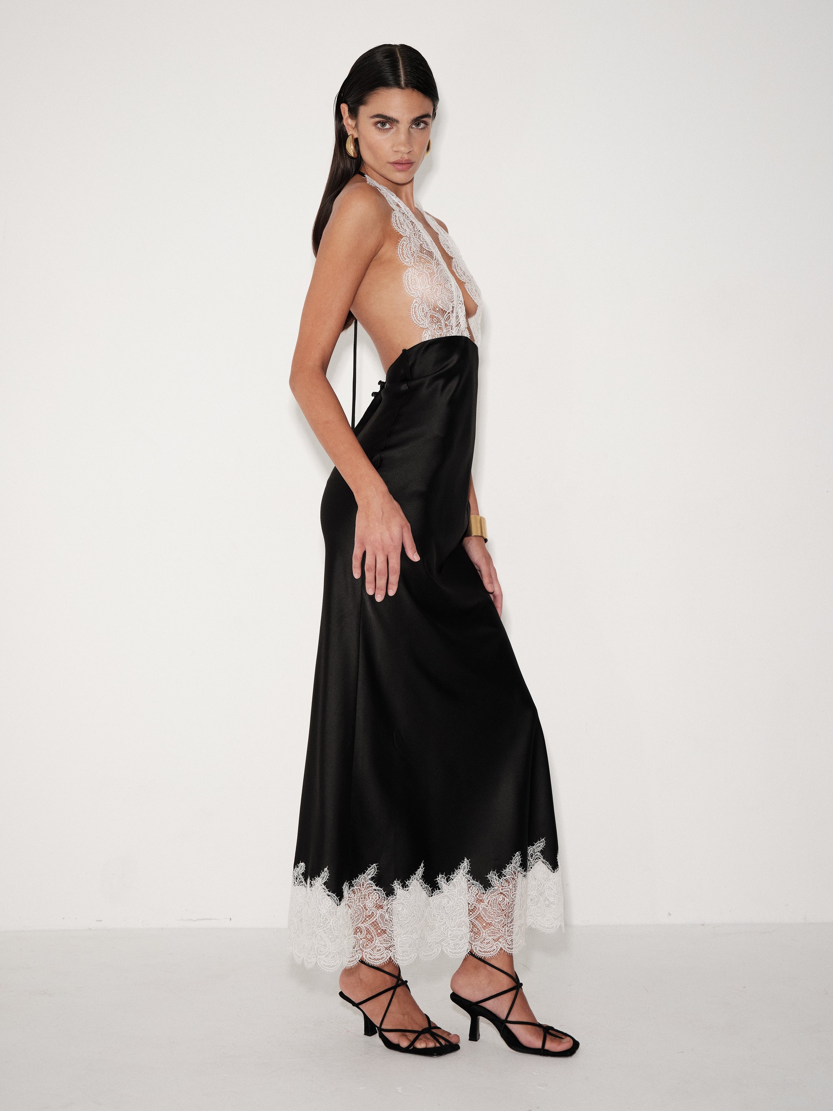 A model wearing a black satin maxi dress with white lace trim and a sheer white lace halter neckline, paired with black strappy high-heeled sandals, set against a plain white studio background.