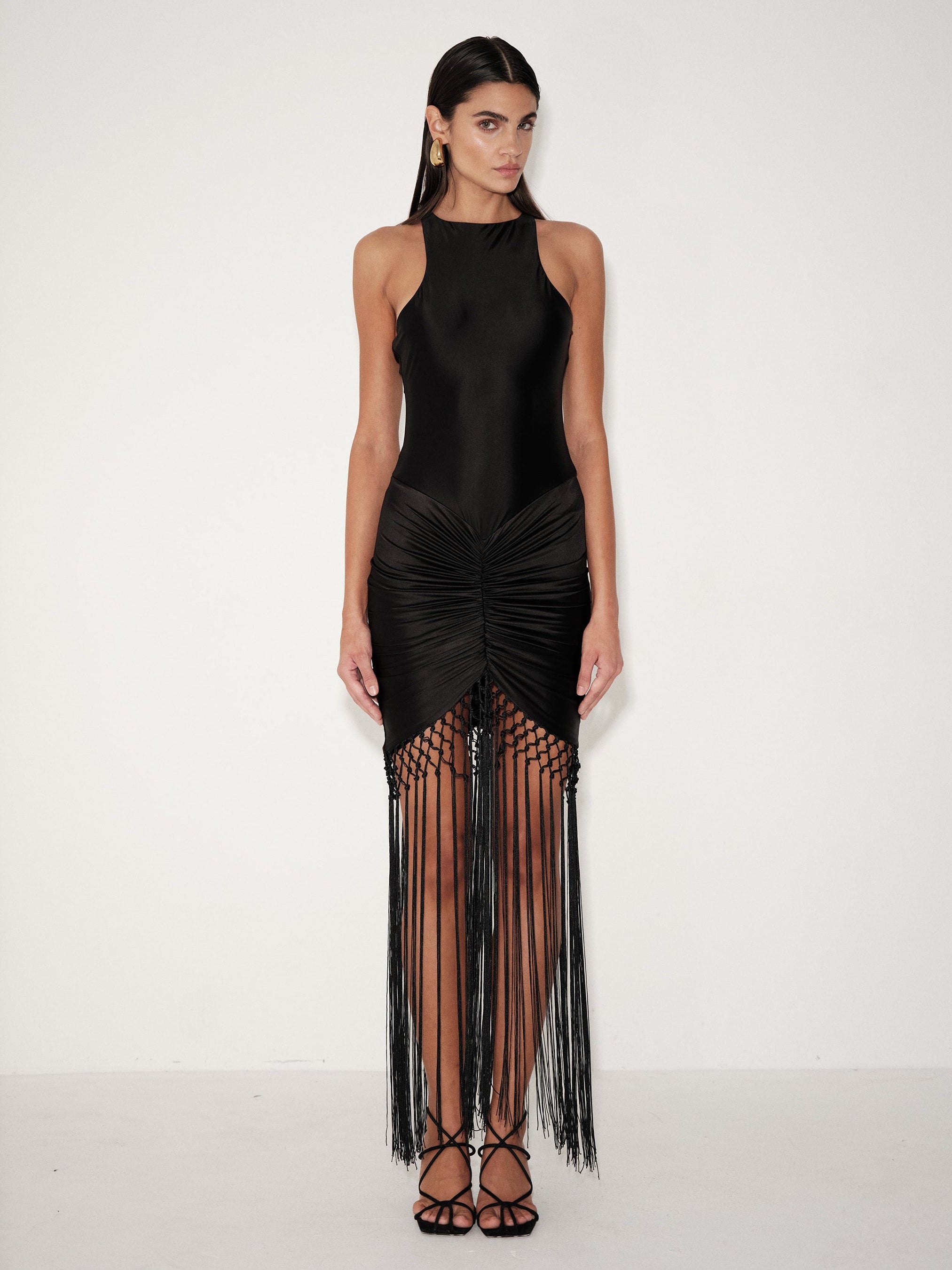 A model wearing a black sleeveless high-neck maxi dress with a ruched gathered skirt and long fringe detailing, paired with black strappy heels, set against a plain white studio background.